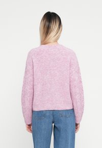 Rosa gestrickter Pullover mit strukturiertem Rücken, im Crewneck-Stil und leicht verkürztem Design. Kombiniert mit blauen High-Waist-Jeans.