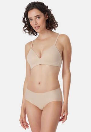Femme aux cheveux bruns bouclés, portant un soutien-gorge beige assorti et des sous-vêtements, debout devant un fond clair uni.