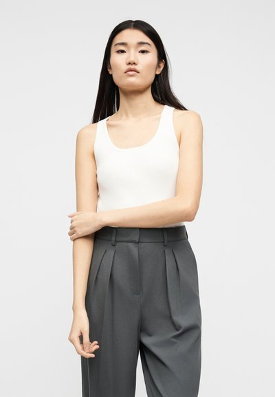 Νεαρή γυναίκα με μακριά σκουρόχρωμα μαλλιά, που φοράει λευκό αμάνικο τοπ και ψηλόμεσα γκρι πιετέ παντελόνια, μπροστά σε απλό φόντο.