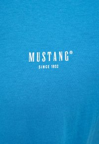 Blue cotton T-shirt featuring a ribbed texture and white print of "MUSTANG" and "SINCE 1932" on the front. Simple, casual design.