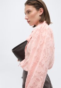 Γυναίκα με κοντά καστανά μαλλιά, που φοράει ροζ υφασμάτινο μπλουζάκι και ασημένια σκουλαρίκια στεφάνια, κρατώντας ένα μαύρο τσαντάκι φάκελο μπροστά σε απλό φόντο.