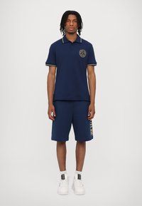 Marineblå polo med gule detaljer og emblemet; kombinert med marineblå shorts med gull "FUTURE" trykk; fullført med hvite joggesko.