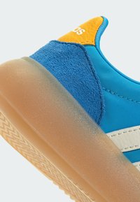 Blue sneaker featuring a combination of smooth fabric, textured suede, and a rubber gum sole. Notable yellow heel accent and white stripes.