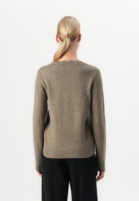 Femeie cu coadă de cal blondă, îmbrăcată cu un pulover tricotat de culoare maro deschis și pantaloni negri, întoarsă cu spatele într-un fundal alb simplu.
