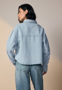 Light blue cropped shirt made of textured fabric, featuring a collared neck, long sleeves, and a back yoke seam. Paired with high-waisted jeans.