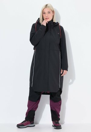 Langer schwarzer wasserdichter Mantel mit Kapuze, ausgestattet mit silbernen Reißverschlüssen und roten Akzenten. Kombiniert mit schwarzen und bordeauxfarbenen Hosen sowie dunklen Wanderschuhen.