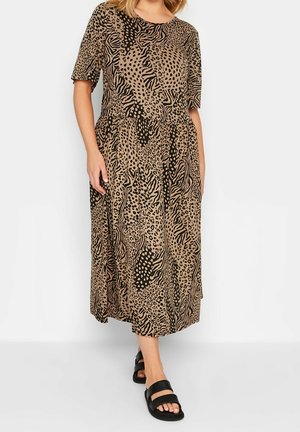 Kvinde iført en knælængde, kortærmet kjole med blandet dyreprint i beige og sort, kombineret med sorte sandaler med åbne tæer.