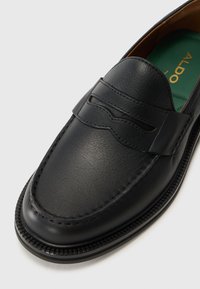 Zwarte leren loafer met een gladde afwerking, voorzien van een gestikt design over de wreef en een ronde neus. Groene binnenzool met logo van het merk.
