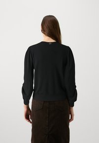 Maglione nero a maglia con maniche a sbuffo, orlo e polsini a coste. Accento dorato sul colletto. Abbinato a una gonna marrone strutturata.
