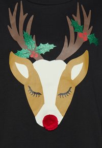 La maglietta nera presenta un design con la testa di un renna, con corna marroni, pelliccia dorata, occhi chiusi e un naso pompon rosso, arricchita da foglie di agrifoglio verdi.