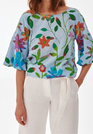 Femme portant un chemisier ample bleu clair avec un imprimé floral coloré et un pantalon blanc taille haute avec une ceinture en tissu nouée à la taille.