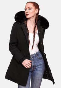 Parka neagră cu un guler mare din blană artificială, închidere cu fermoar și manșete cu dantelă. Purtați-o peste un top alb și blugi albaștri, având o siluetă subțire.