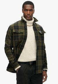 Chaqueta de camisa a cuadros verdes y negros con bolsillos en el pecho, botones negros y un cuello alto, superpuesta sobre un suéter de cuello alto crema.