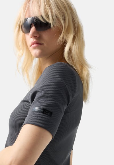 Lyshåret kvinde iført sorte solbriller og en grå ribstrikket kortærmet top med "FIRE + ICE" mærke på ærmet, set fra siden.