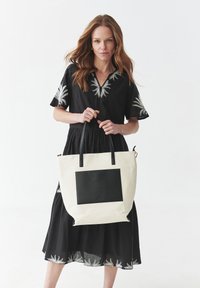 Torba typu tote z płótna w kolorze kremowym, z czarną skórzaną kieszenią z przodu; posiada czarne uchwyty i przestronny design. Elementy dekoracyjne nie są wliczone.