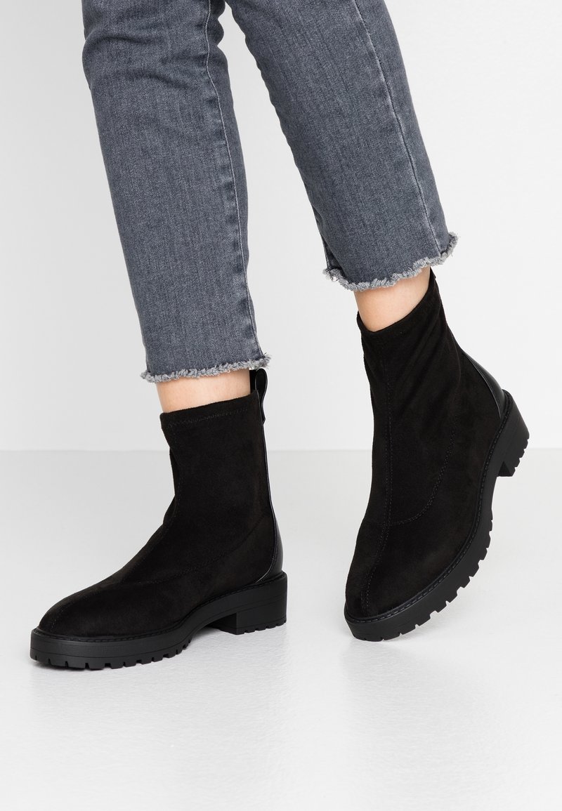 Bottines en daim noir à finition lisse, dotées d'une semelle épaisse et d'une languette à l'arrière, associées à un jean gris à ourlet effiloché.