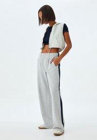 Lichtgrijze, cropped zip-up vest met capuchon over een marineblauwe top, gecombineerd met grijze sweatpants met marineblauwe zijkanten en een trekkoord in de taille.