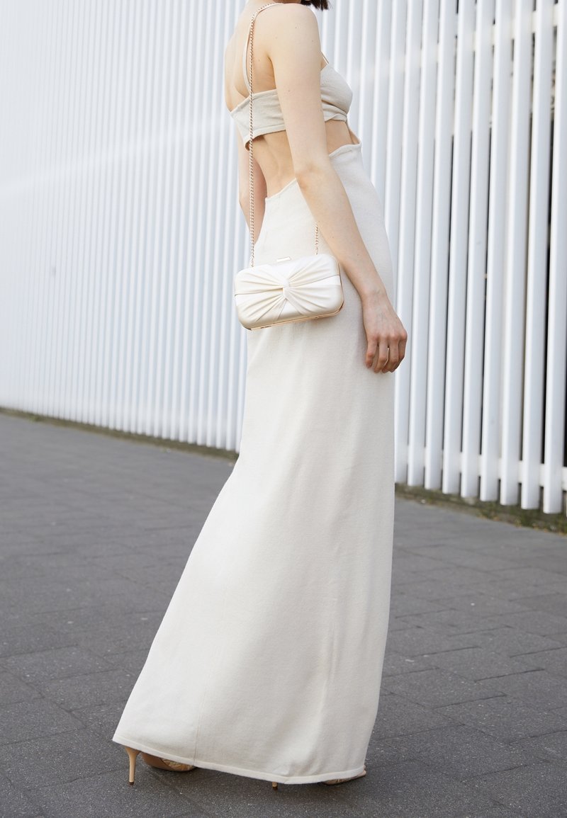 Femme portant une robe beige dos nu avec de fines bretelles, tenant une petite pochette assortie avec une bandoulière chaîne, debout sur un trottoir pavé.