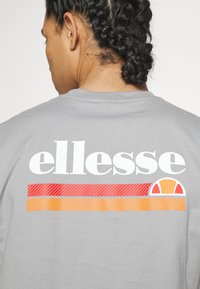 Gray cotton t-shirt with a large white "ellesse" logo and horizontal stripes in red and orange. Features a round logo accent.