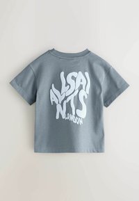 Harmaa puuvillapaita lyhyillä hiekoilla, jossa on suuri, vääristynyt valkoinen "ALLSAINTS LONDON" -grafiikka takana.