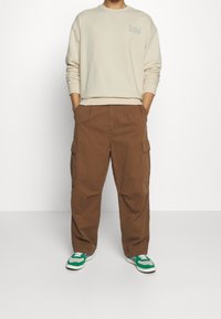 Krémový svetr s kulatým výstřihem a logem "Lee", doplněný hnědými cargo kalhotami se bočními kapsami a zelenými teniskami.