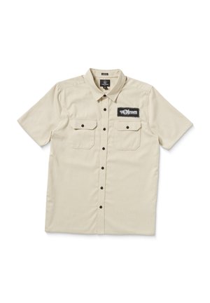 Short-sleeve beige button-up shirt with a black logo patch, two chest pockets, and black buttons, featuring a textured, woven fabric.