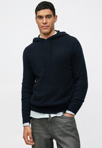 Tamsiai mėlyna džemperis su raišteliu, raglaniniais rankovėmis ir minkšta tekstūra, uždressuotas ant šviesiai mėlynos juostuotos marškinėlių ir pilkų džinsų.