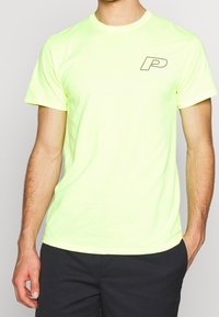 Neonově žluté bavlněné tričko s kulatým výstřihem a krátkými rukávy, se černým logem "P" na levé části hrudi. Hladká textura.