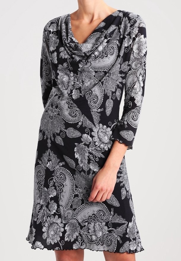Kobieta w czarnej sukience z długimi rękawami, ozdobionej białymi wzorami paisley i kwiatowymi, z dekoltem w kształcie drapowania i falbaniastym dołem.