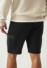 Shorts noirs à texture lisse, dotés d'une poche latérale zippée et d'un petit logo sur la cuisse gauche, associés à un sweat-shirt beige.