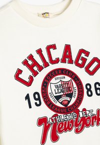Sudadera blanca con cuello acanalado que presenta estampados en rojo y azul marino. El texto incluye "CHICAGO" y "Athletic Dept. New York" con un diseño de escudo.