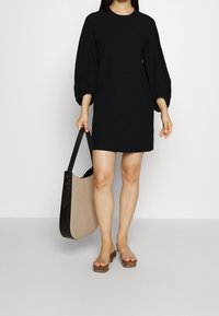 Robe noire mi-longue avec des manches bouffantes, faite d'un tissu lisse. Accompagnée d'un sac fourre-tout beige et noir et de sandales à talons transparents.