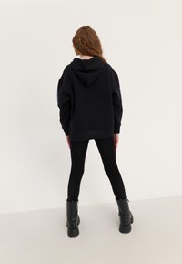 Hoodie oversized preto com capuz, tecido texturizado e detalhes impressos nas mangas. Usado sobre leggings pretas e botas pretas robustas.