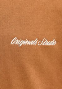 Krátky rukáv, hnedá tkanina s hladkou textúrou featuring biele, písané vyšívanie, ktoré znie "Originals Studio."