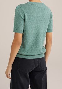 Groene gebreide trui met korte mouwen, een textuurpatroon, ronde hals en geribbelde boorden en zoom, gedragen met zwarte high-waisted broek.