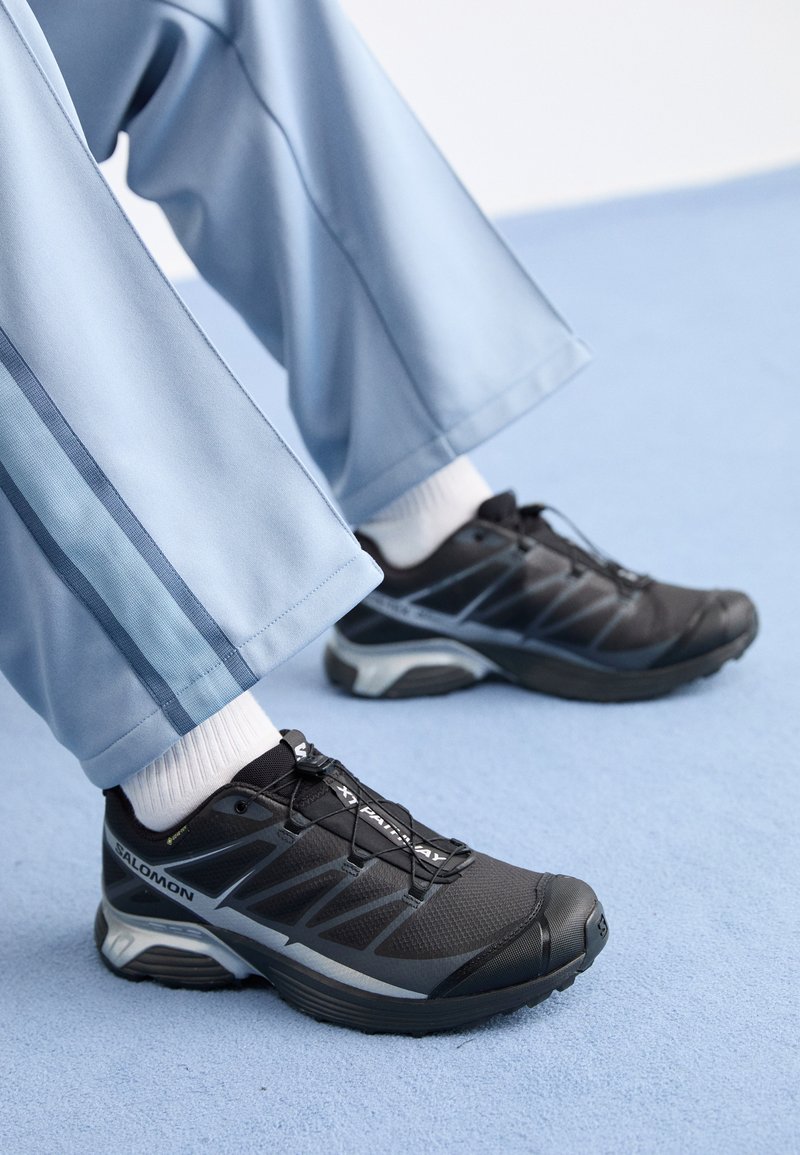 Black athletic shoes with a textured upper, reflective accents, and a thick sole, paired with light blue track pants and white socks.