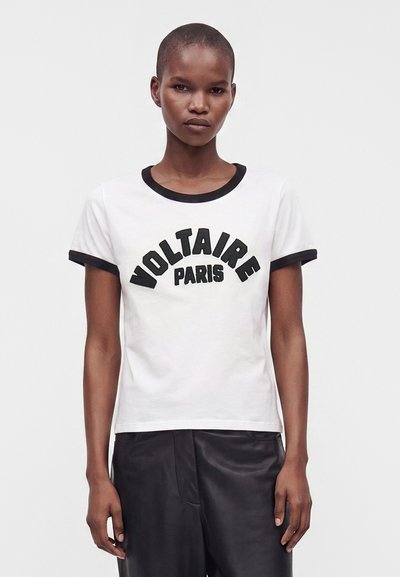 Μοντέλο με κοντά μαλλιά που φοράει λευκό T-shirt με μαύρο γιακά και μανίκια, μαύρο κείμενο "Voltaire Paris," και μαύρο παντελόνι.