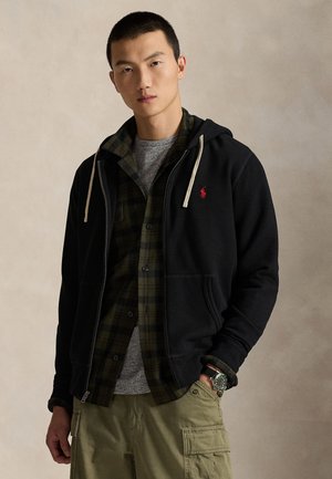 Ung mand iført en sort lynlåshættetrøje over en grøn ternet skjorte, grå t-shirt, beige cargobukser og et armbåndsur, står mod en neutral baggrund.