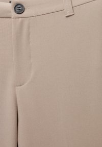 Beige nadrág gombos záródással, övhurkokkal és elülső zsebbel, közeli nézetben látható szövettextúrával.