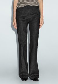 Vrouw in een hooggelegde, donkergrijze, uitlopende broek met een zwart-wit gestreept bovenstuk dat erin is gestopt, staat tegen een eenvoudige lichte achtergrond.