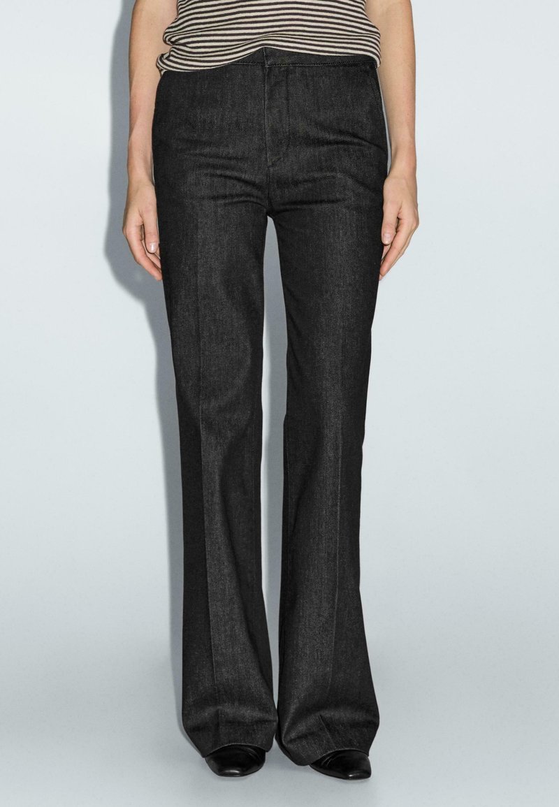 Vrouw in een hooggelegde, donkergrijze, uitlopende broek met een zwart-wit gestreept bovenstuk dat erin is gestopt, staat tegen een eenvoudige lichte achtergrond.