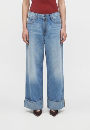 Persona che indossa jeans azzurri a gamba larga con grandi risvolti, abbinati a scarpe scure con punta a punta e top rosso senza maniche.