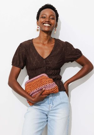 Femme souriante aux cheveux courts et bouclés portant un haut marron à col en V et un jean bleu clair, tenant une pochette orange et rose crochetée.