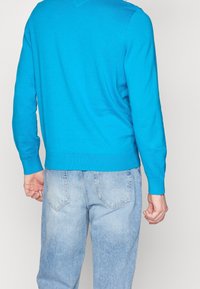 Pull en tricot bleu à manches longues, avec poignets et ourlet côtelés. Associé à un jean en denim bleu clair présentant une finition délavée et des poches arrière.