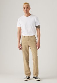 Beige katoenen broeken met een rechte pijp, gecombineerd met een wit T-shirt met ronde hals. Model draagt zwarte sneakers met witte zolen.