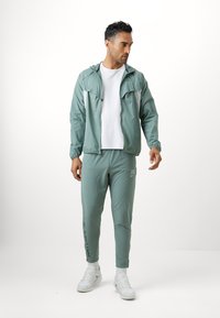 Světle zelená bunda se zipem vpředu, kontrastní bílé panely, odpovídající světle zelené tepláky s textem na boku a bílé tenisky.