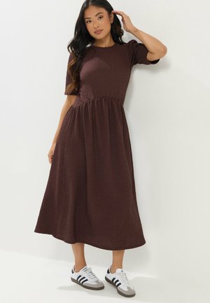 Frau in einem strukturierten dunkelbraunen Midikleid mit kurzen Ärmeln, stehend und sich an den Haaren berührend, trägt weiße Adidas-Sneaker mit schwarzen Streifen.