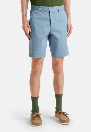 Hellblaue Baumwollshorts mit flachem Bund, normalem Taillenbund und umgeschlagenen Säumen. Getragen mit grünen Socken und beigen Bootsschuhen.