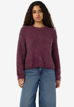 Jeune femme aux longs cheveux bouclés portant un pull violet duveteux et un jean bleu, debout devant un fond blanc uni.
