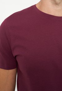 Nahaufnahme einer Person, die ein schlichtes kurzärmeliges kastanienbraunes T-Shirt mit rundem Ausschnitt vor hellem Hintergrund trägt.